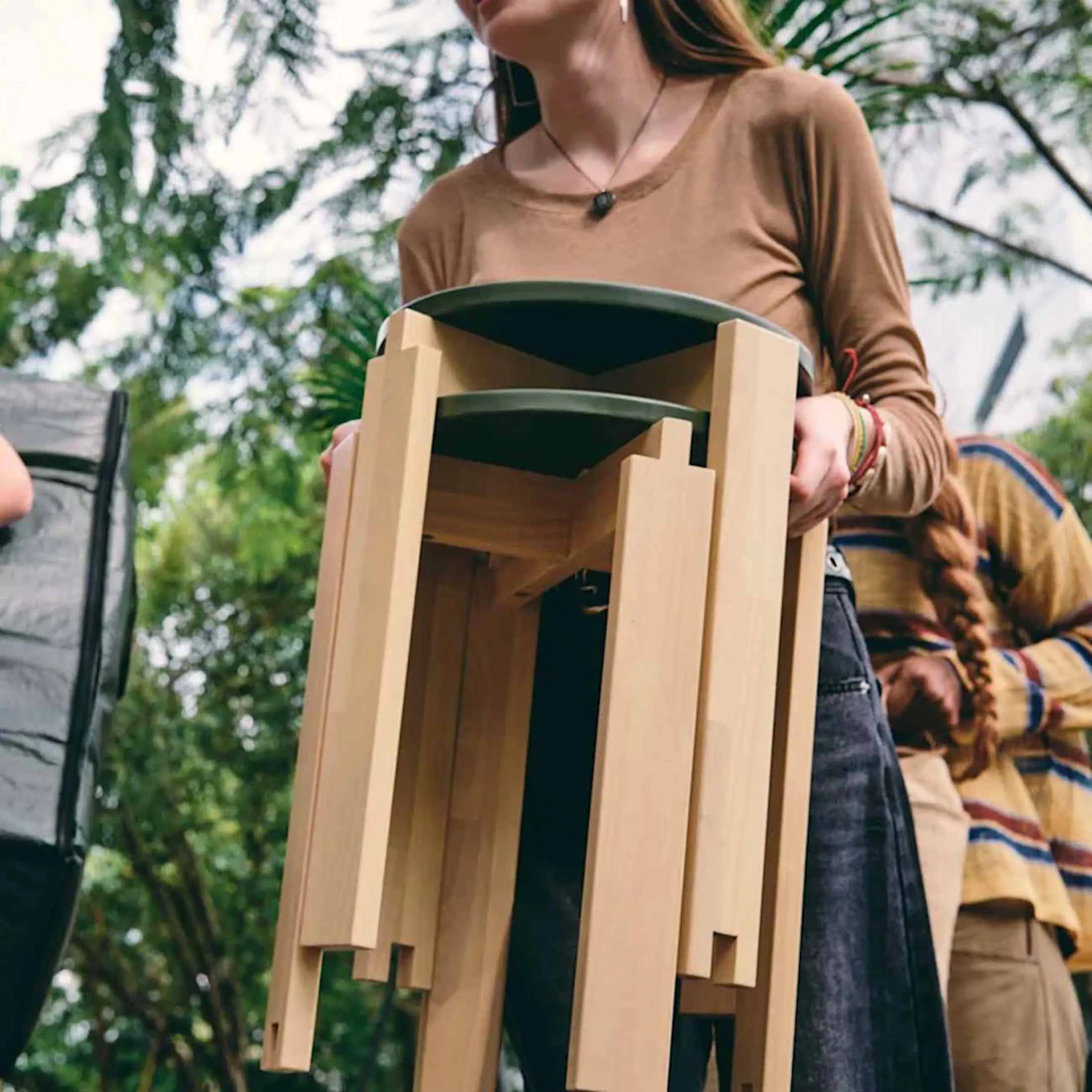 A person carrying two stacked IKEA KOMPISHÄNG stools outdoors, the notched birch legs visible from below