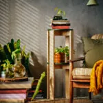 Two IKEA KOMPISHÄNG stools interlocked into a two-tier pedestal, with a potted plant on the lower green seat and books and a glass vessel on the upper one