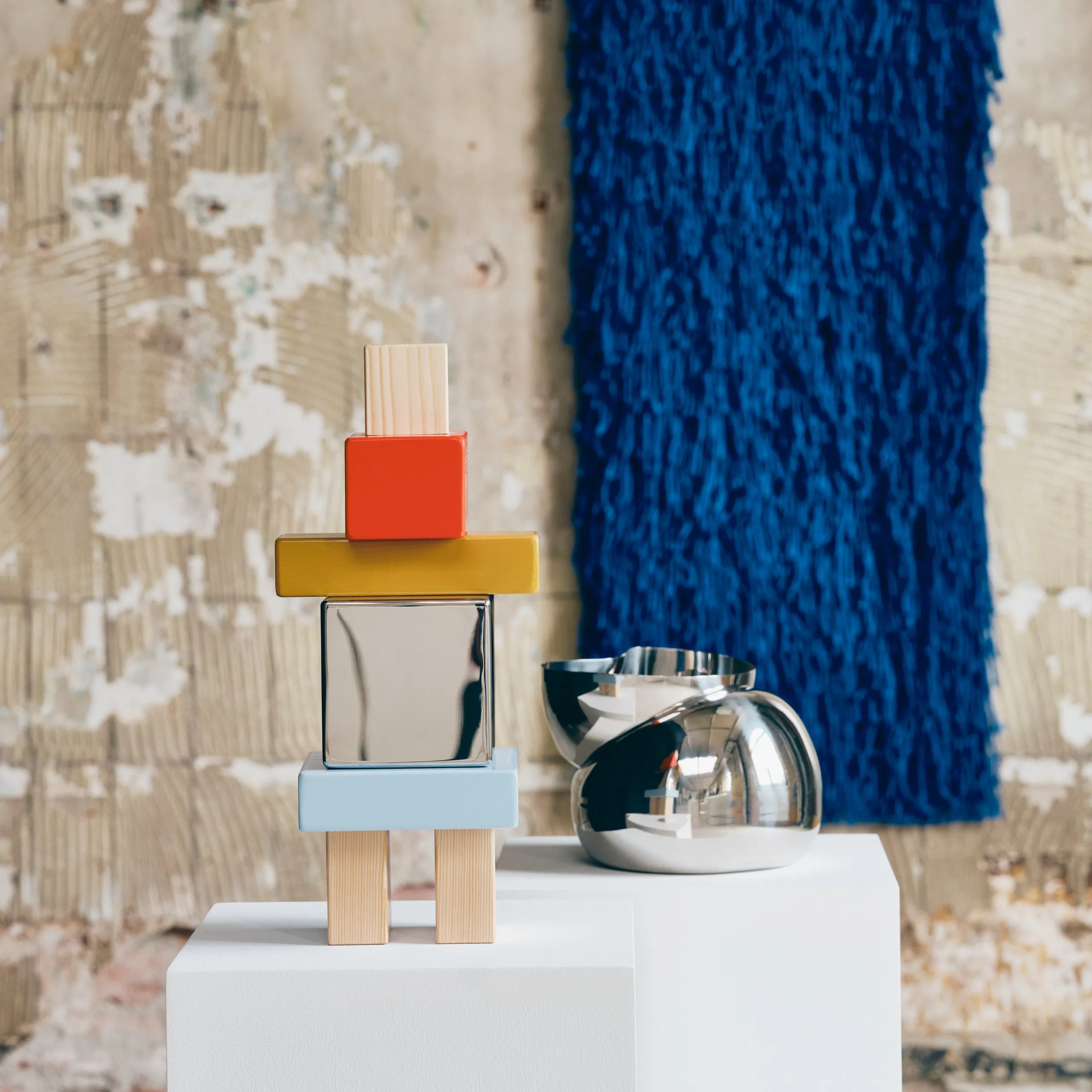 Colorful KONSTRUNDA guardian sculpture and stainless-steel bowl with a blue textile behind them
